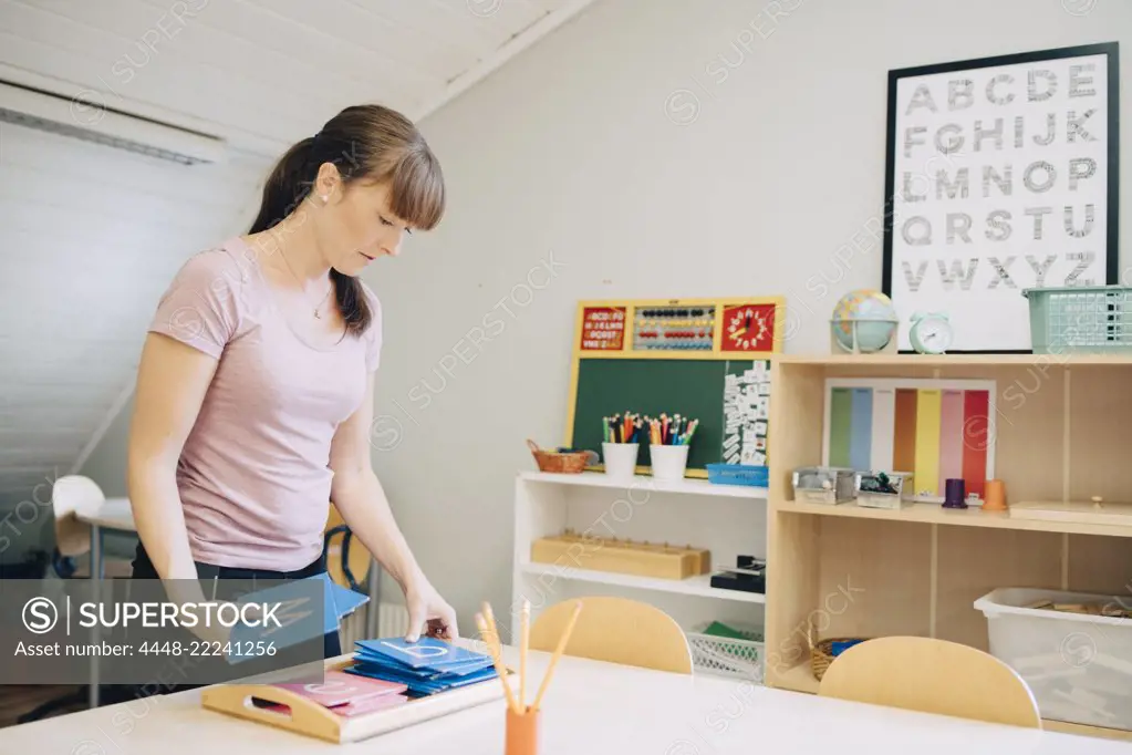 Confident mid adult female teacher arranging flash cards with alphabets at table in classroom