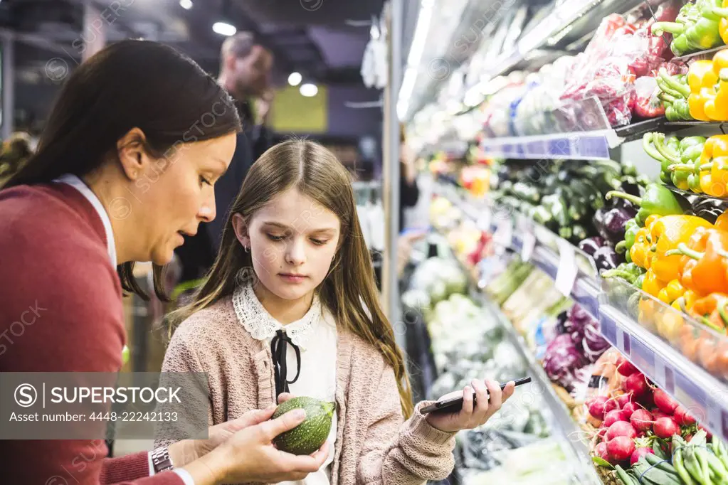 Mother showing cucumber to daughter while grocery shopping in supermarket