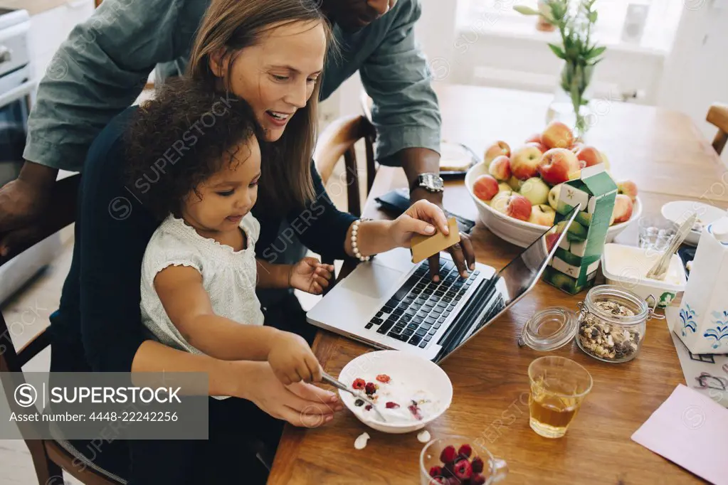 Parents shopping online on laptop while looking at daughter having breakfast in dining room