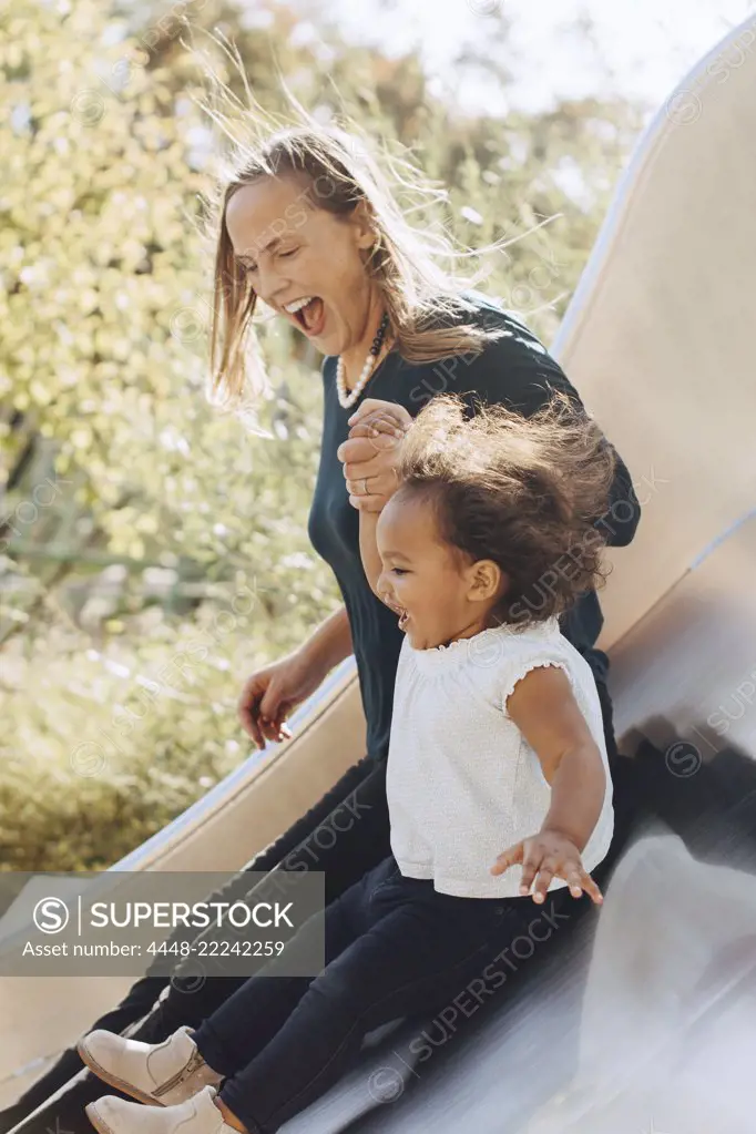 Happy mother sliding with daughter on slide at park