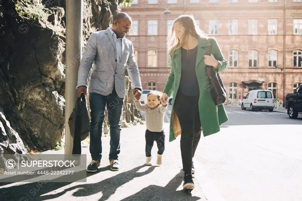 Full length of multi-ethnic parents holding daughter's hands while walking on footpath at city