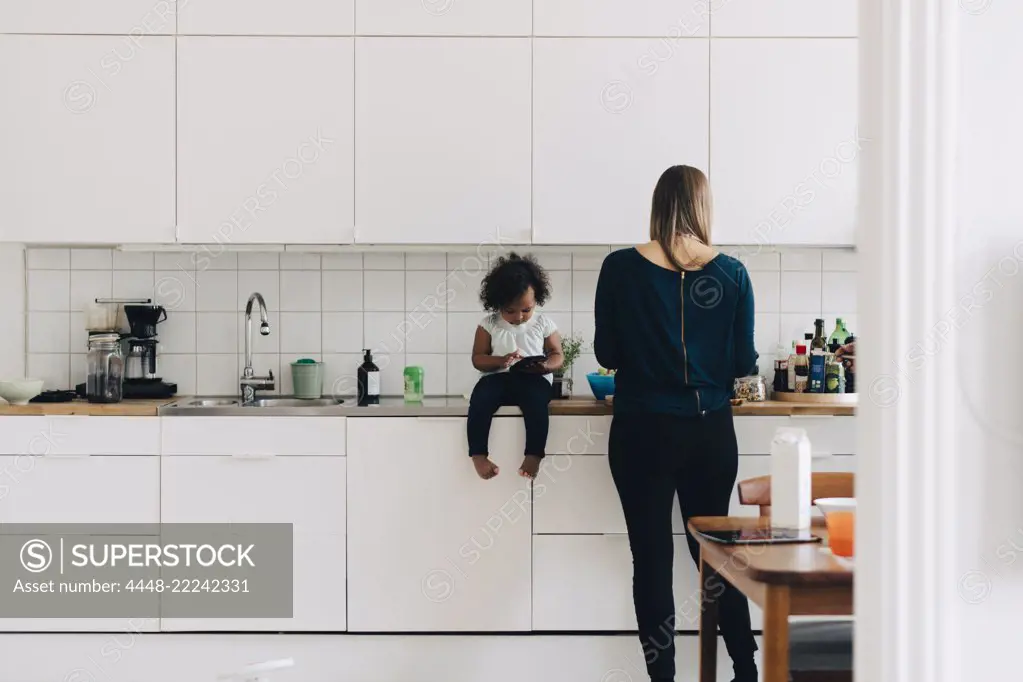 Full length of girl using smart phone while mother working in kitchen at home