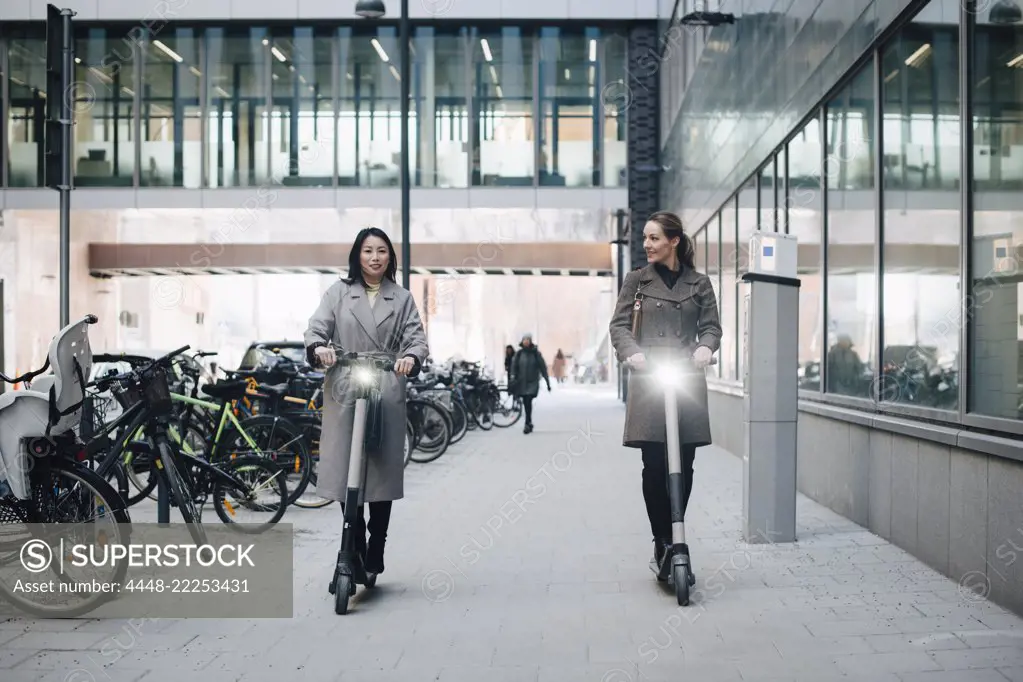 Full length of confident female colleagues riding electric push scooter on footpath by building in city