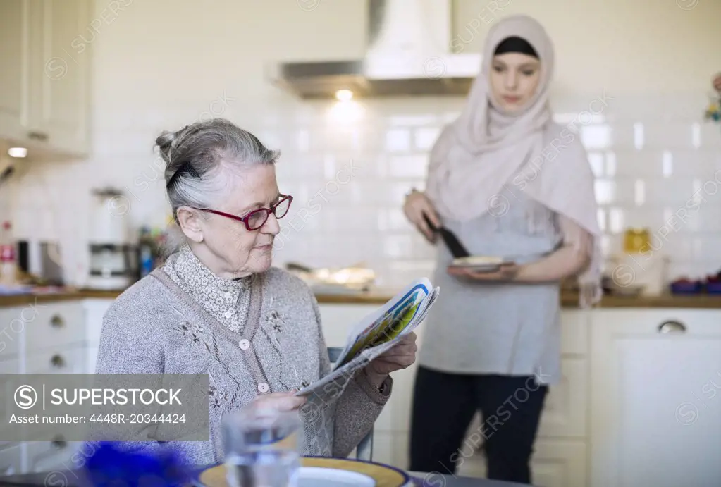 Senior woman reading newspaper with female home caregiver preparing food in kitchen