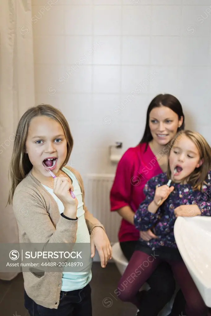 Portrait of girl brushing teeth with mother and daughter in bathroom