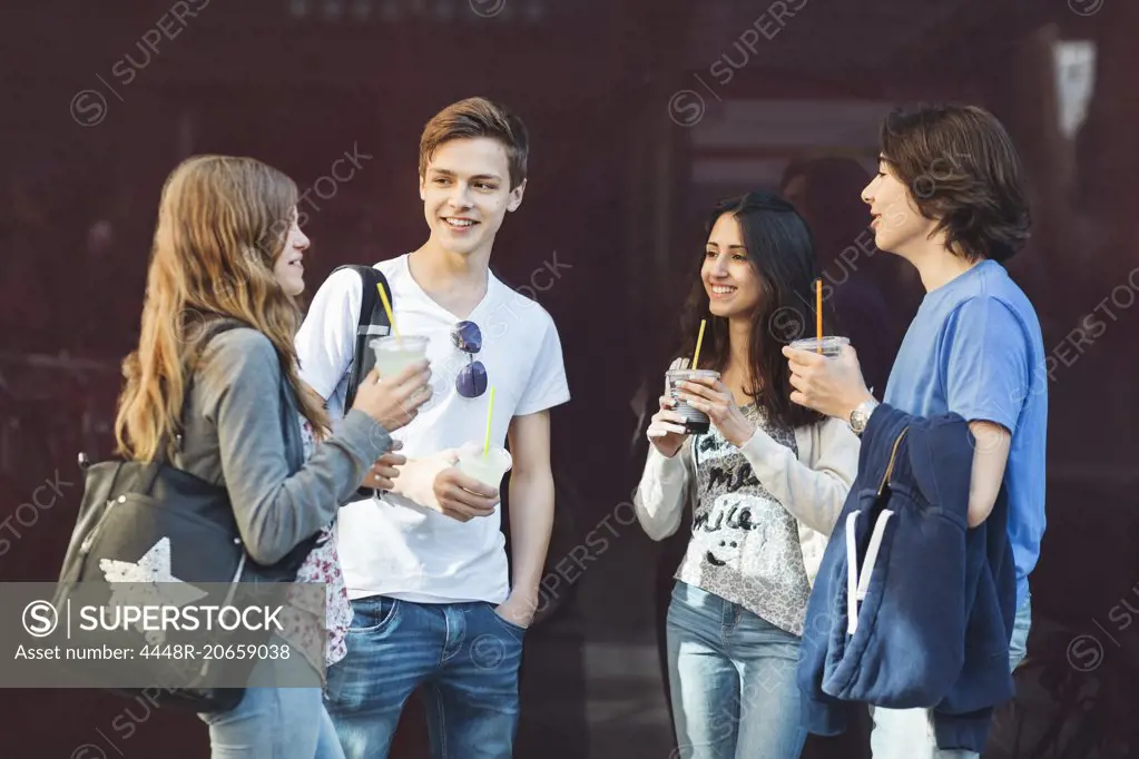 Happy teenage friends talking while holding drinks outdoors