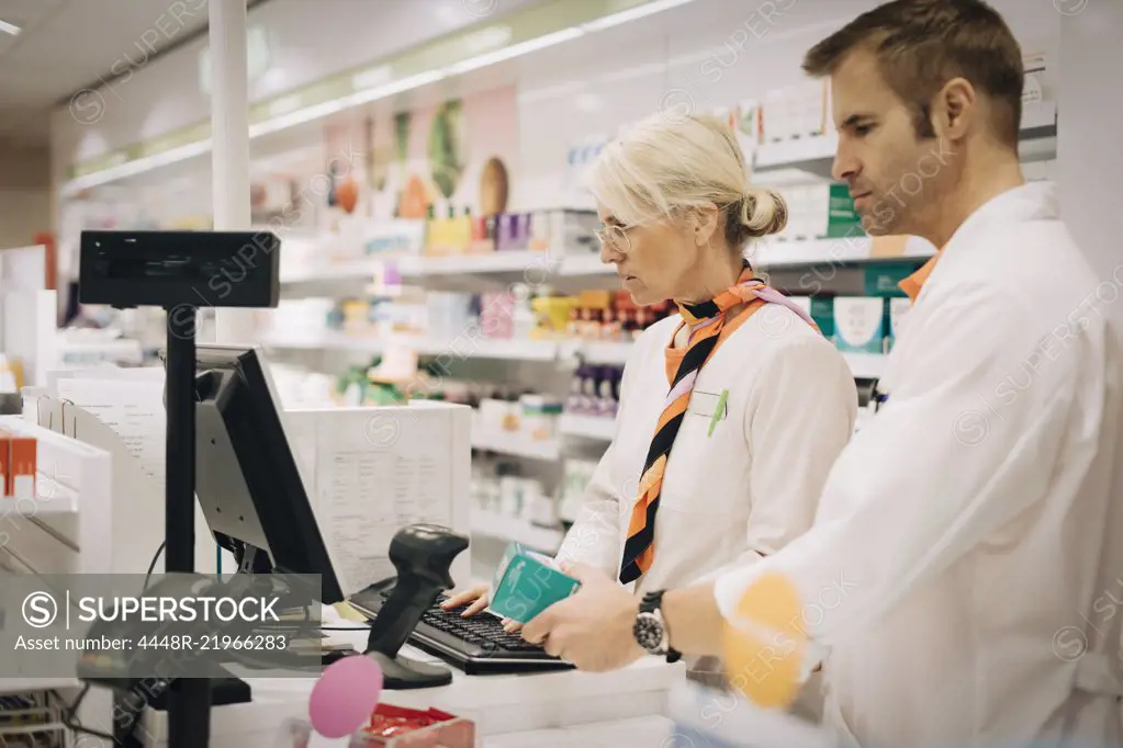 Confident male pharmacist scanning medicine with bar code reader by female colleague using computer at checkout in store