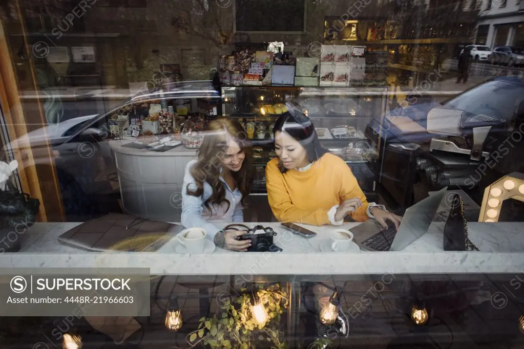 Female bloggers with camera and laptop sitting at cafe seen through window glass