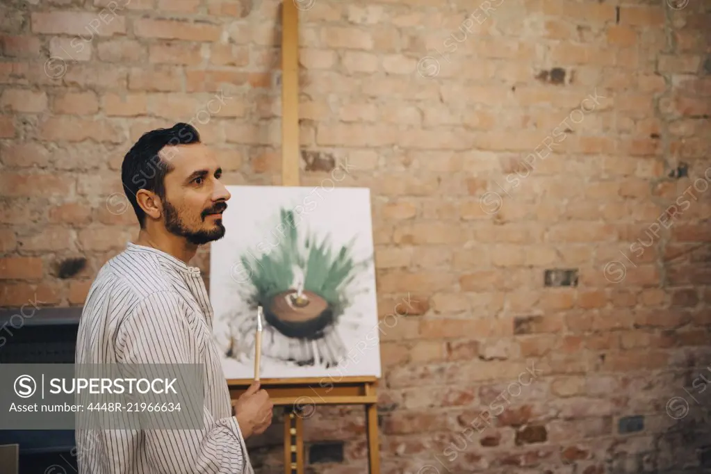 Side view of male artist standing by painting against brick wall at studio