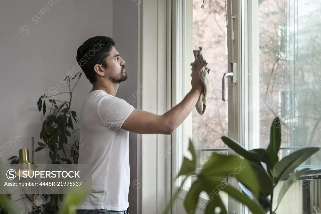 Side view of young man cleaning glass window in living room