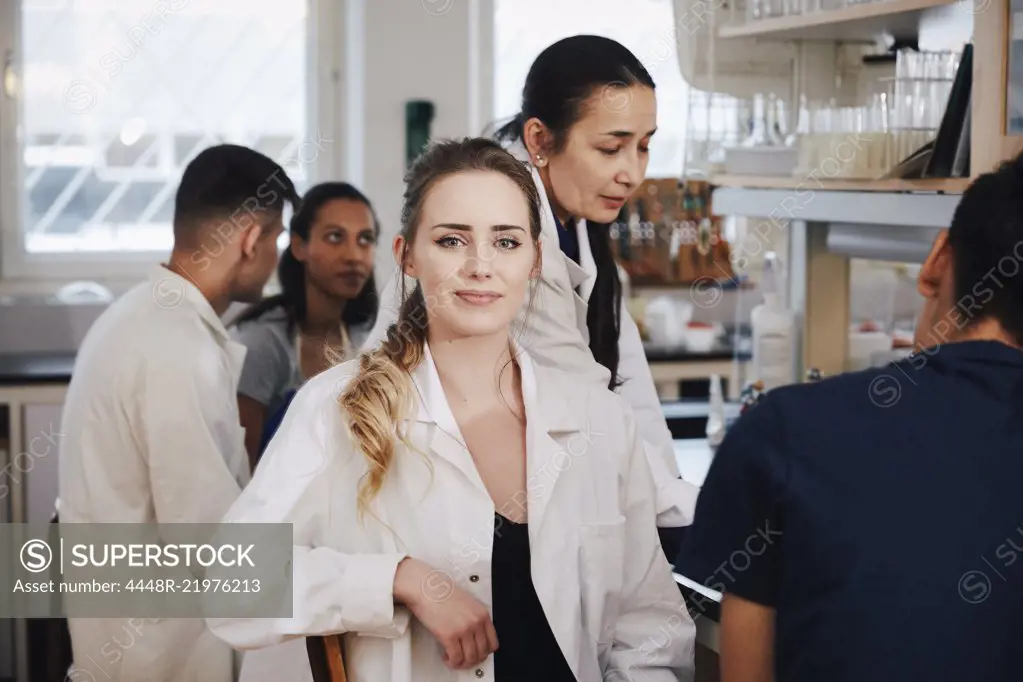 Portrait of confident young female student sitting against teacher friends in chemistry laboratory