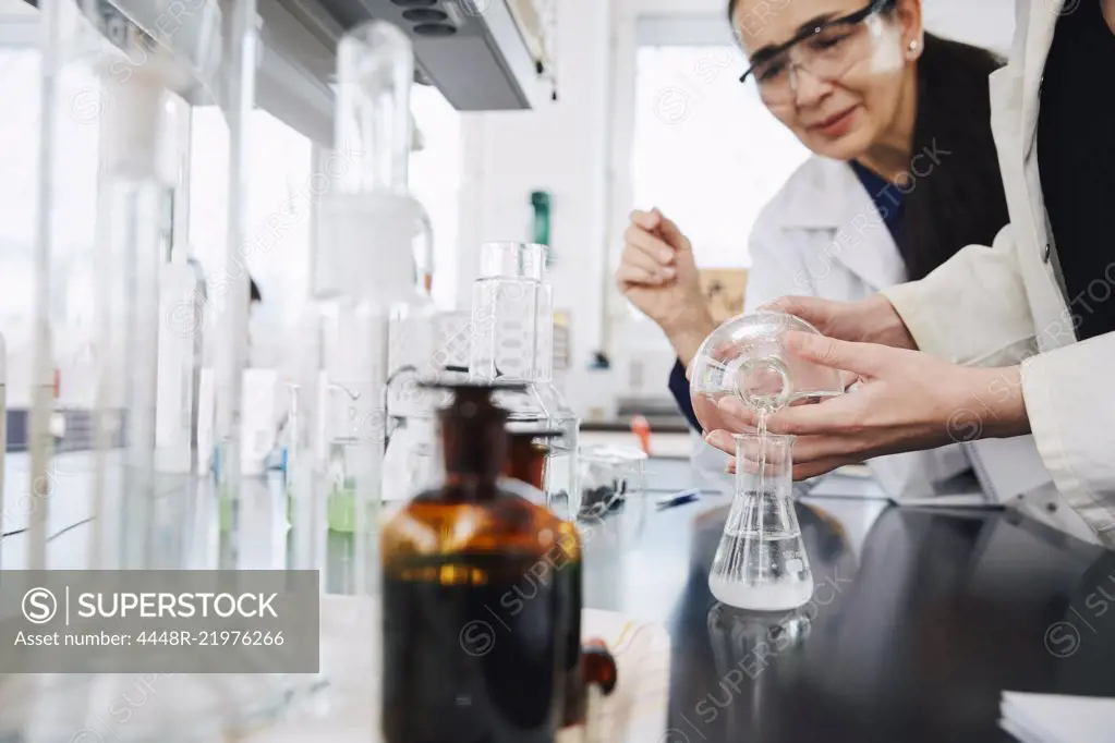 Midsection of young female student pouring solution in flask by teacher at chemistry laboratory