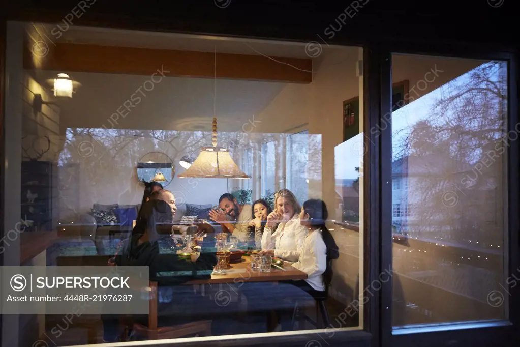 Happy family having dinner at table seen through glass window during sunset