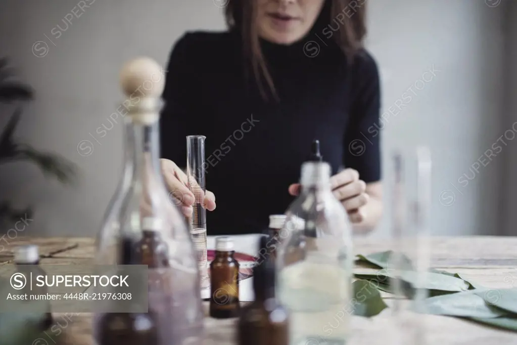 Midsection of female owner mixing liquid while sitting at table against wall in perfume workshop