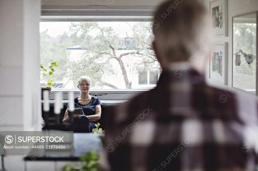 Rear view of senior man looking at woman sitting on sofa in living room