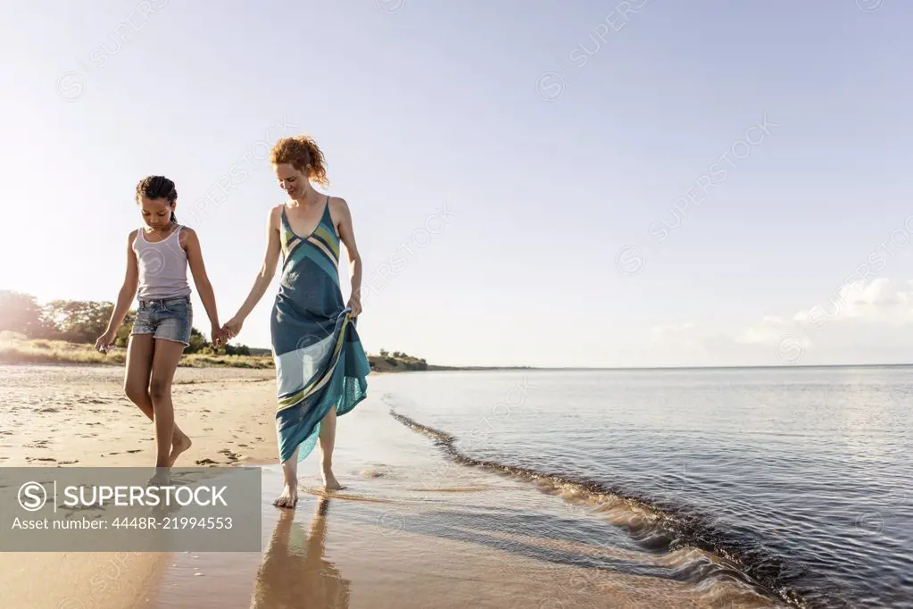 Full length of woman holding hands with daughter while walking on shore at beach against sky