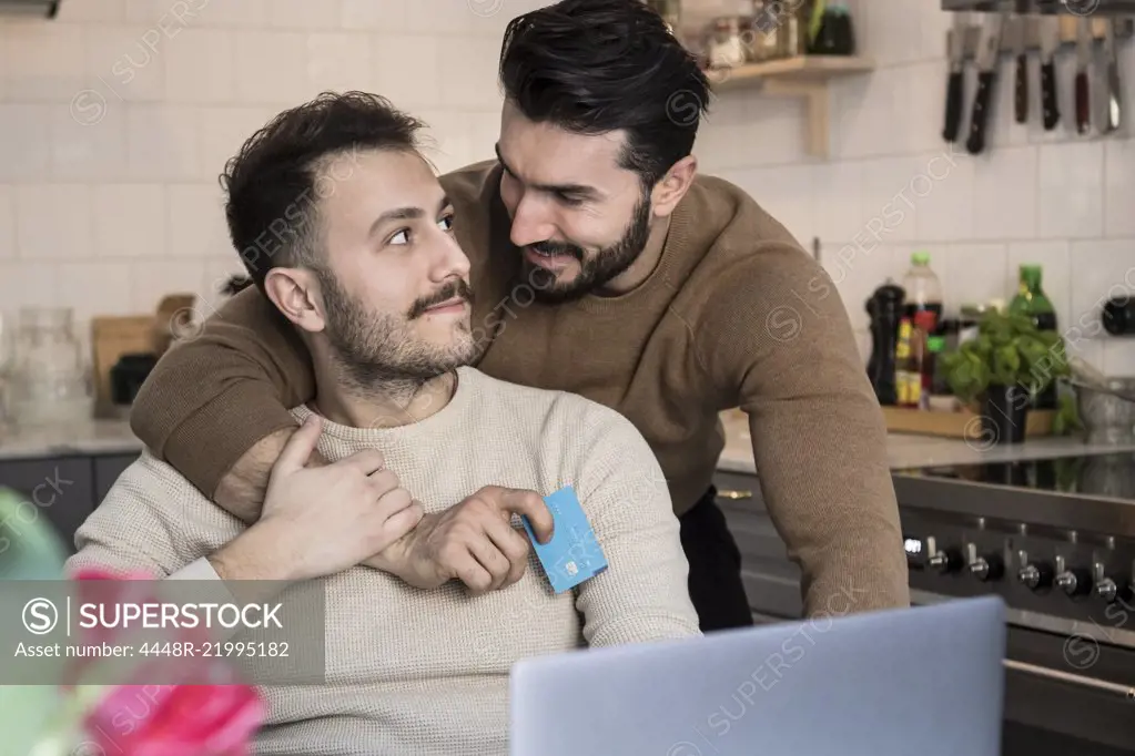 Romantic gay couple doing online shopping through laptop in kitchen at home