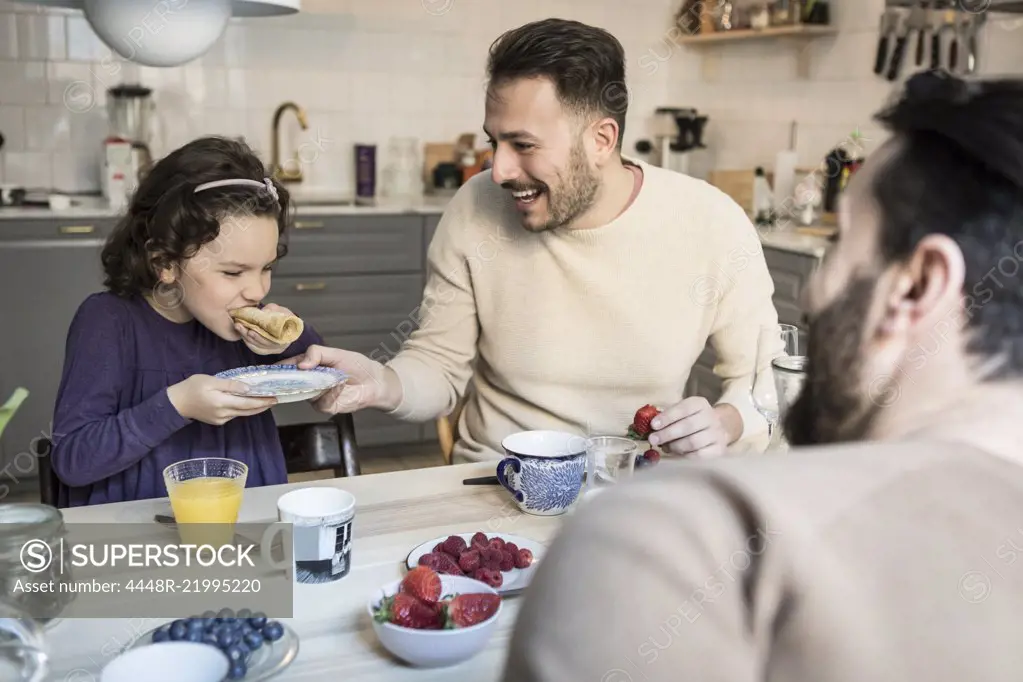 Laughing father holding plate while daughter eating pancake at table