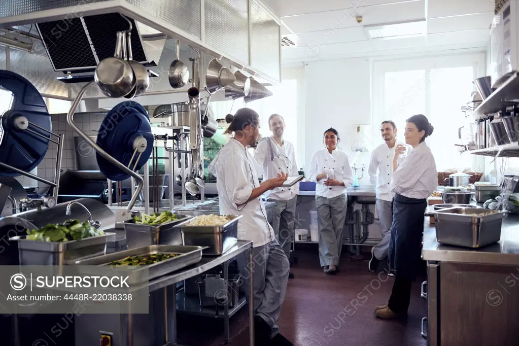 Male chef holding digital tablet while discussing with colleagues in commercial kitchen