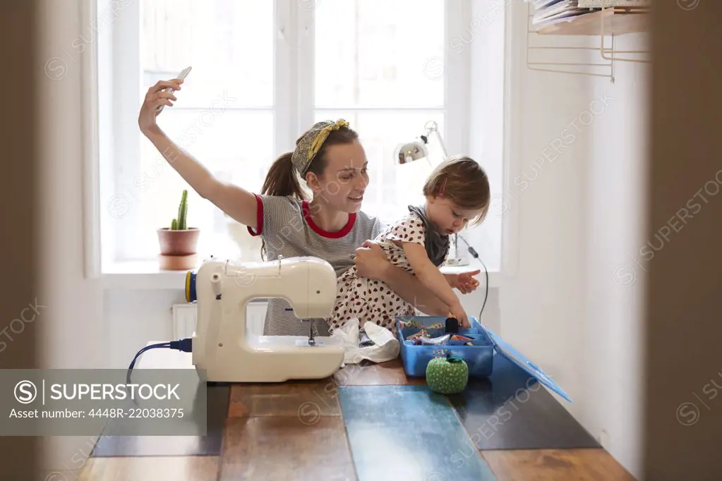 Female tailor taking selfie with daughter through smart phone while sitting on table at home