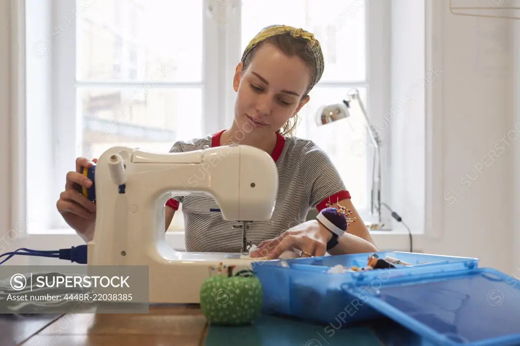 Young tailor using sewing machine while sitting against window at home