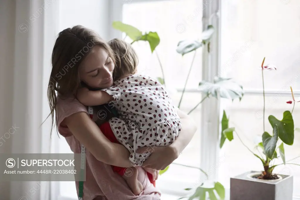 Female fashion designer embracing daughter while standing by window at home