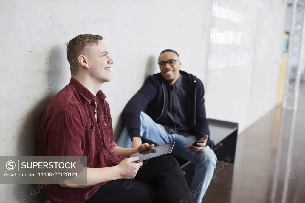 Smiling student holding digital tablet while sitting with friend on bench in university