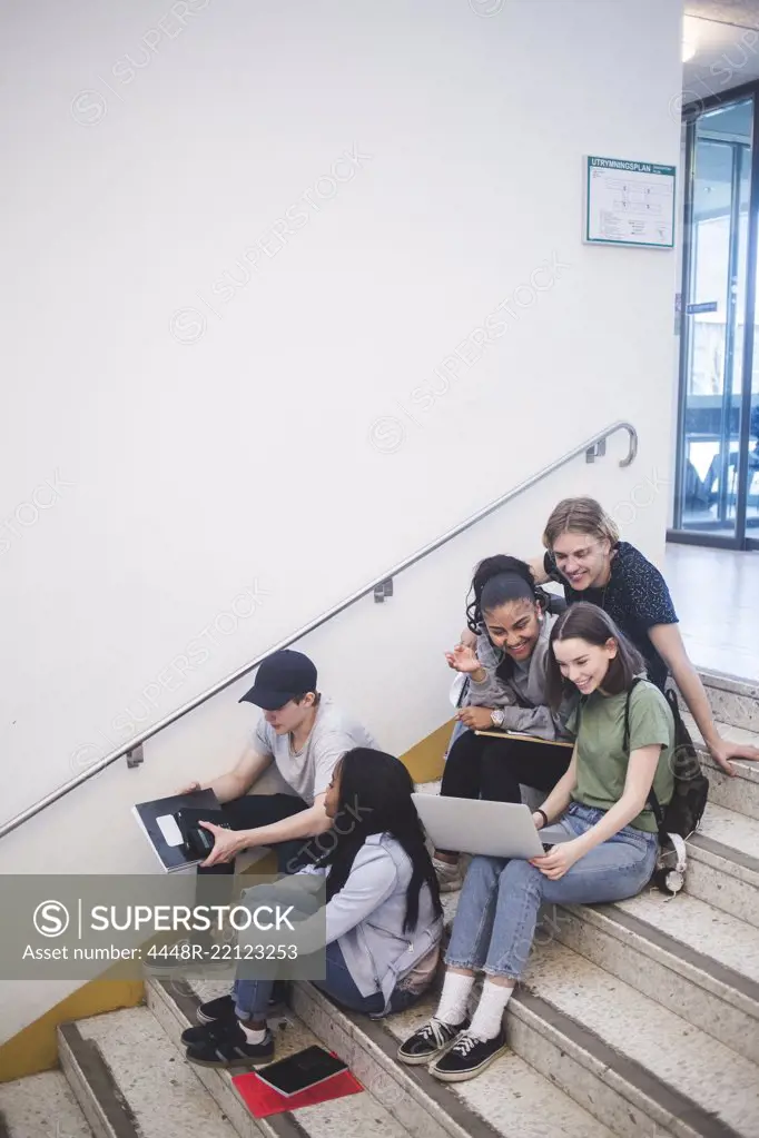 Full length of happy multi-ethnic students talking and sitting on steps at high school