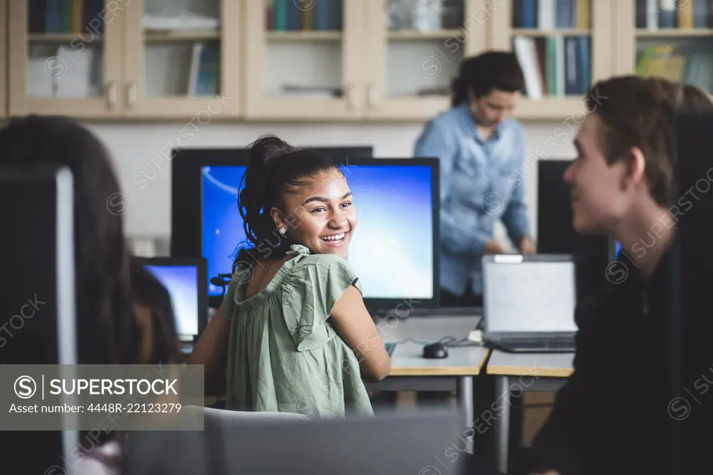 Smiling female high school student looking at friend sitting in computer lab