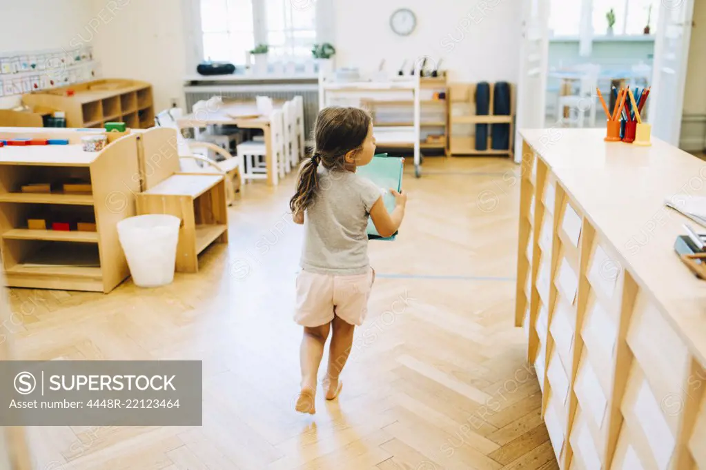 Full length rear view of girl running with book in classroom at kindergarten