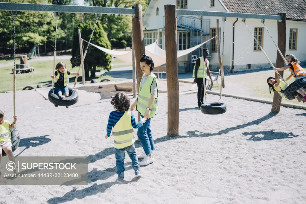 Teachers standing while students enjoying on swing in playground