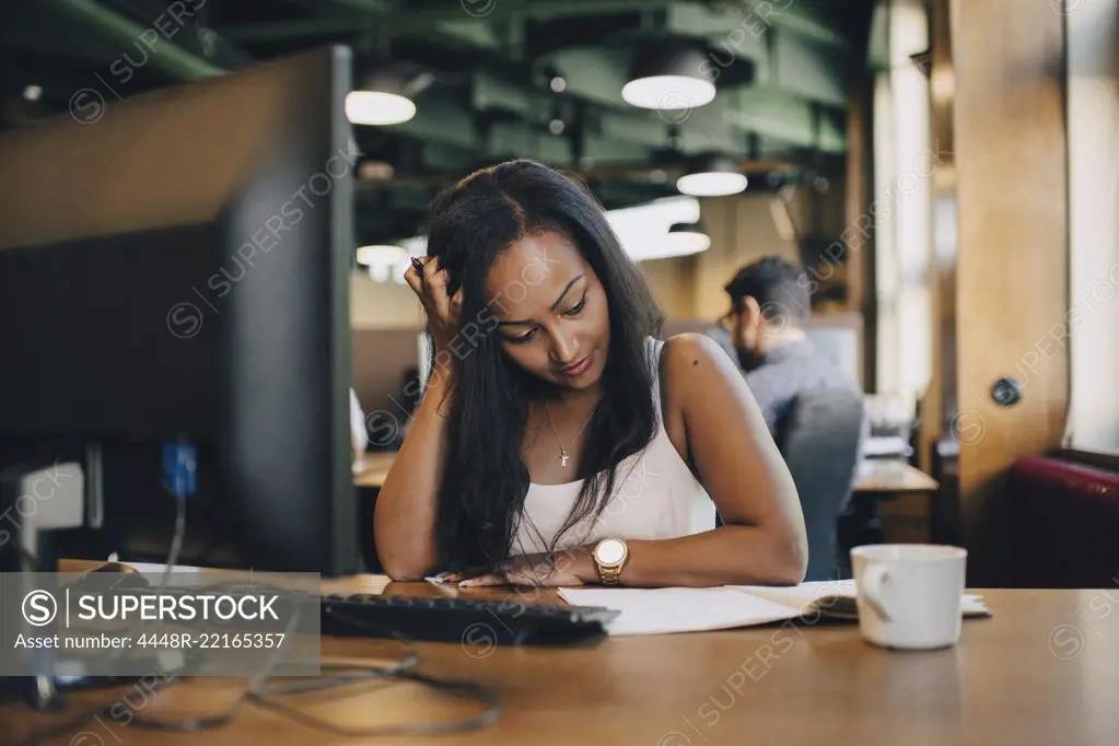 Businesswoman with hand in hair reading book at desk in office