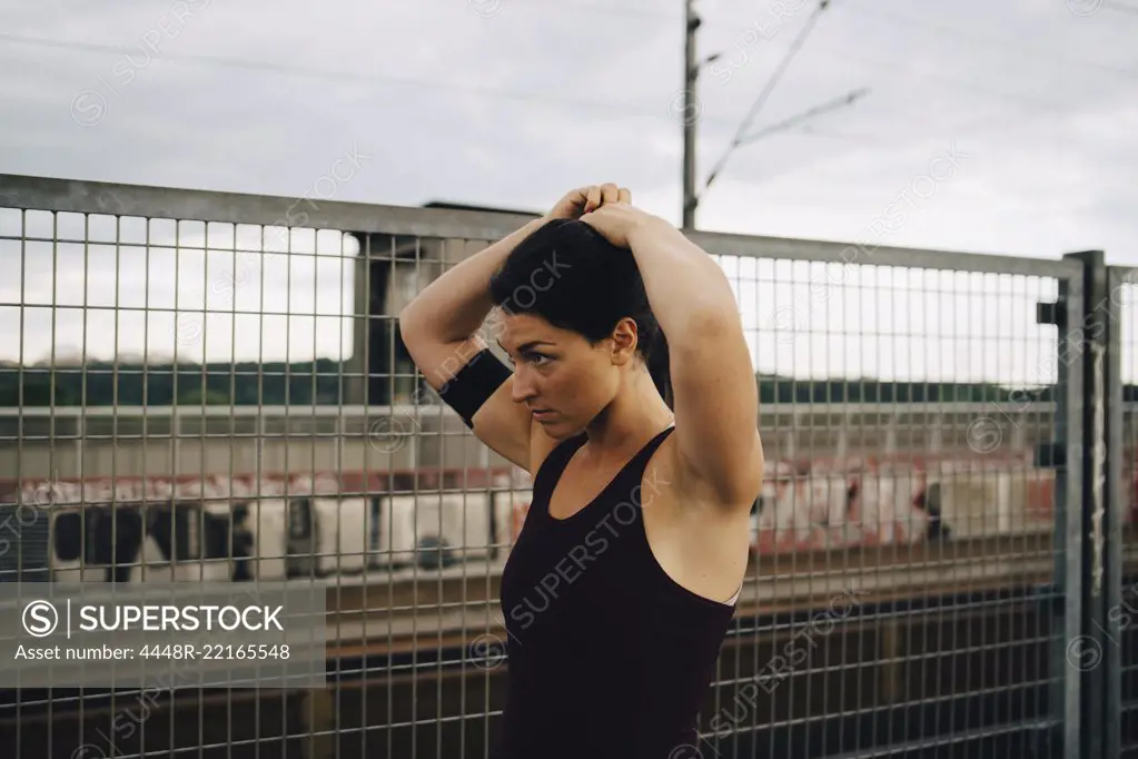 Determined female athlete tying hair while standing by fence on footbridge