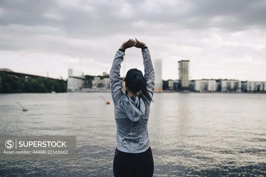 Rear view of female athlete stretching arms while standing against sea in city