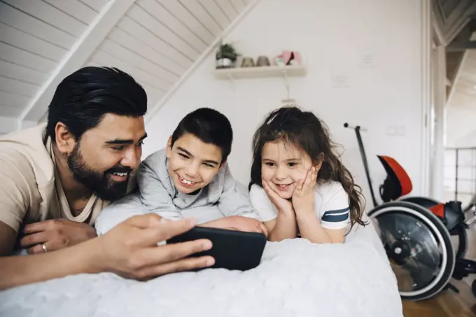 Smiling father showing video to children over mobile phone while lying on bed at home