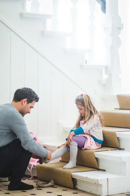 Father assisting schoolgirl wearing shoes while sitting on steps at home