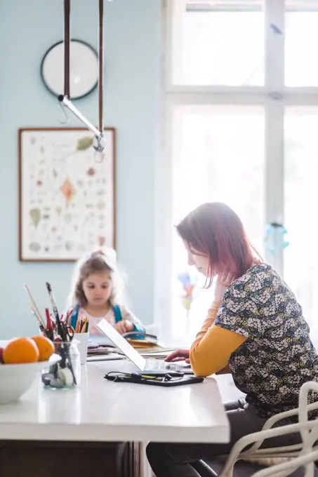 Side view of mid adult women working on laptop while sitting with girl at kitchen island