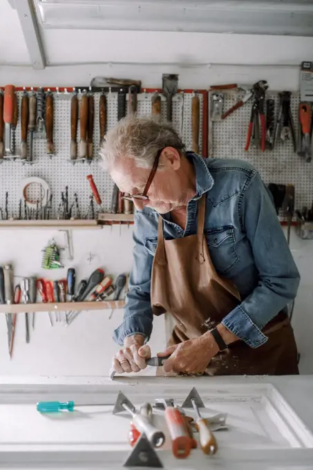 Senior male entrepreneur scraping window frame on workbench at store workshop