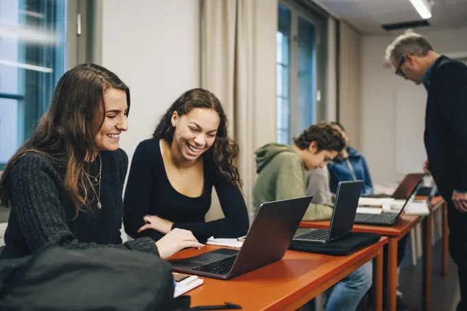 Cheerful female high school students using laptop at desk in classroom