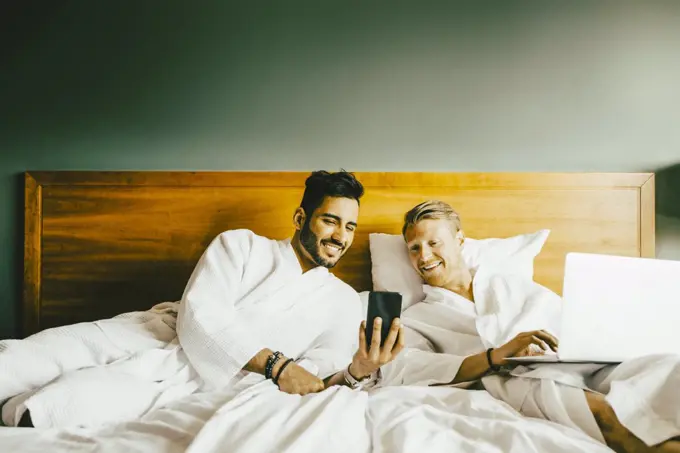 Smiling man showing smart phone to boyfriend using laptop while leaning in bed at hotel