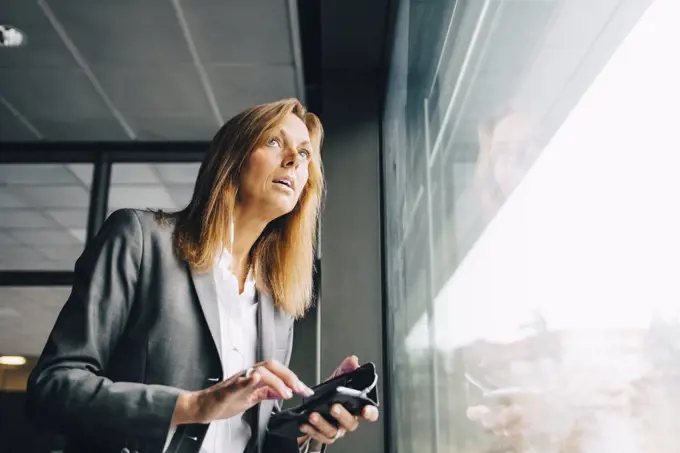 Thoughtful businesswoman with mobile phone looking through window in office