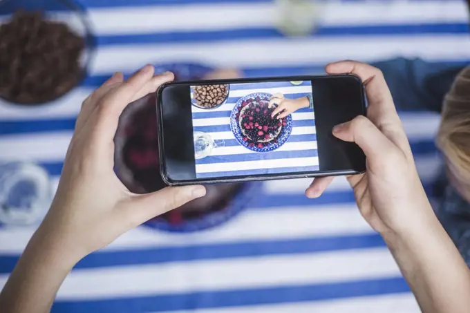 Cropped hand of girl with smart phone photographing boy having dessert at table