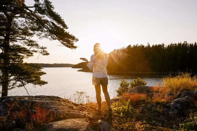 Rear view of woman holding mobile phone while standing on lakeshore at sunset