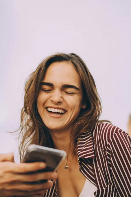 Cheerful young woman looking at smart phone held by friend at terrace