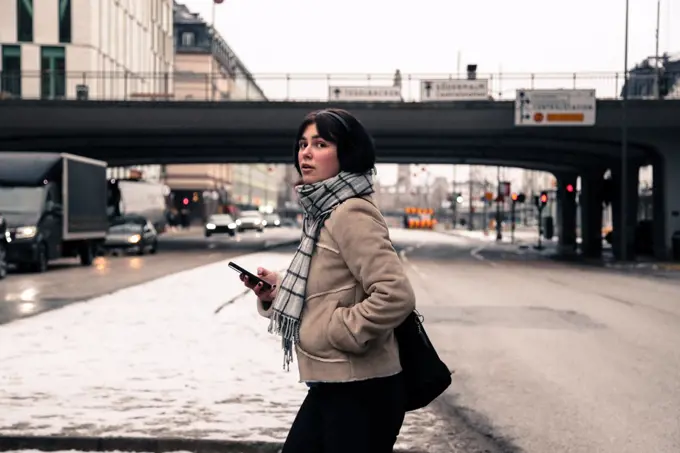 Side view of young woman holding mobile phone while crossing road in city during winter