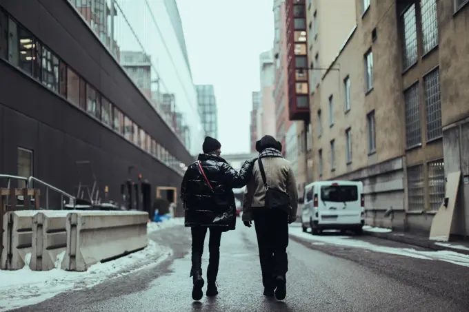 Full length rear view of female friends wearing warm clothing while walking on wet street in city by buildings