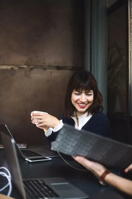 Smiling female entrepreneur holding coffee cup discussing with colleague over document at desk in office