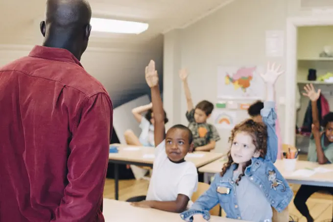 Students raising hand while looking at teacher in classroom at school