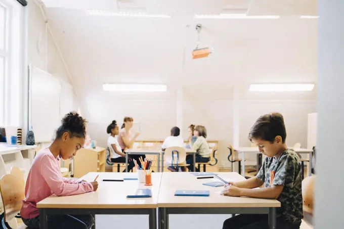 Boy and girl writing at desk while friends learning with student in background