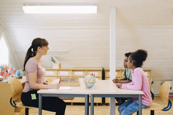 Teacher talking to school boys while sitting at desk in classroom